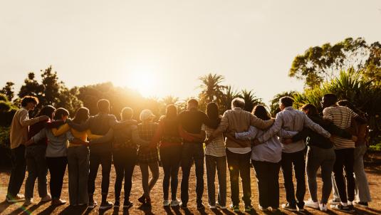 Stock image of a group of people shoulder-to-shoulder, backs facing the camera and looking up towards the sun.