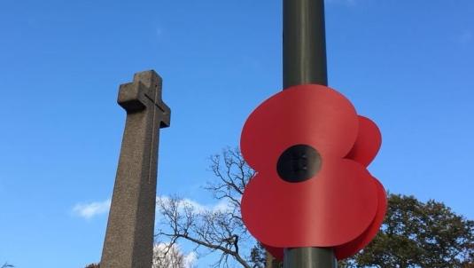 LArge poppy attached to lamppost in foreground, tall war memorial cross in background with blue sky