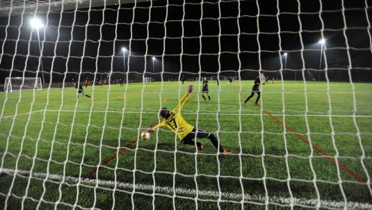 Floodlit view from behind a goal of a football pitch with goalkeeper diving for the ball