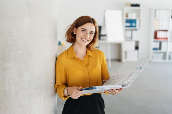 A young woman in professional business attire, holding a folder and smiling at the camera.