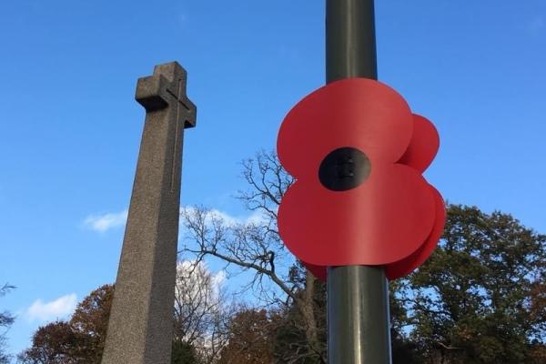 LArge poppy attached to lamppost in foreground, tall war memorial cross in background with blue sky