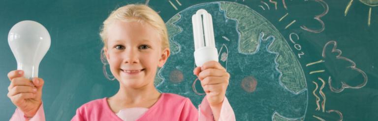 [A child holding up an old fashioned lightbulb and an energy efficient lightbulb. They are standing in front of a blackboard with a drawing of Earth on it]