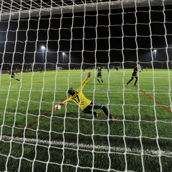 Floodlit view from behind a goal of a football pitch with goalkeeper diving for the ball